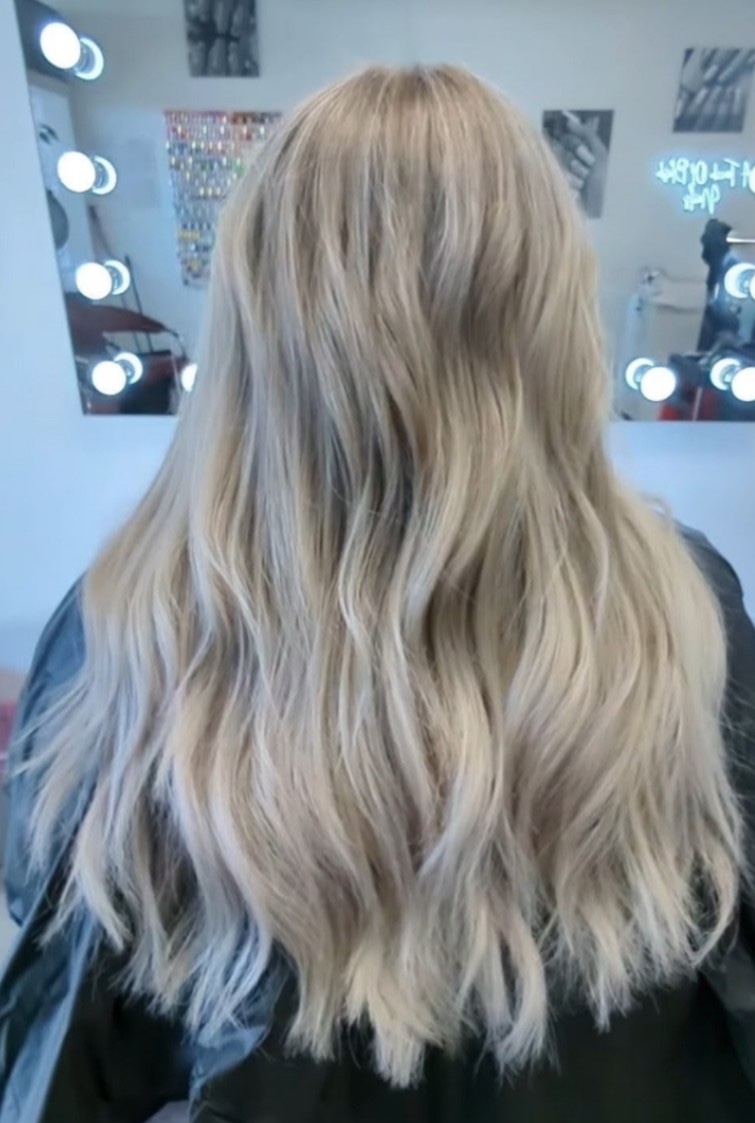 Back view of long, soft blonde waves in front of a lit vanity mirror at the studio